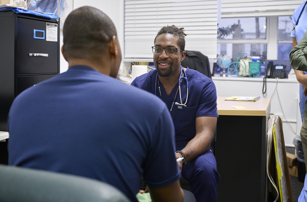 A member of Penn Medicine’s United Community Clinic at the African Family Health Organization sits in an office chair, smiling, in front of a patient.  